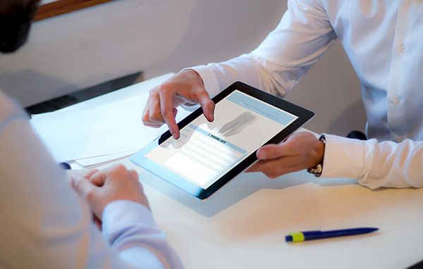 A person is holding a tablet, and another person is gesturing towards it while seated across the table.