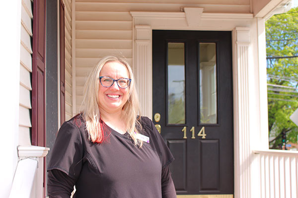 The image shows a woman standing in front of a house, smiling at the camera.