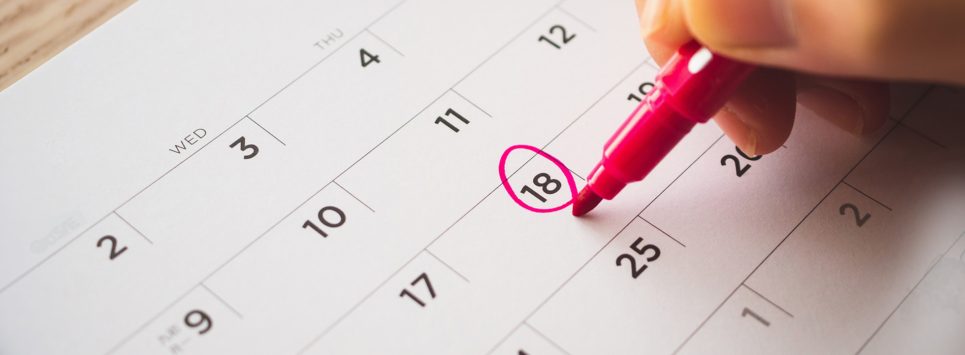 The image shows a close-up of a hand holding a pink highlighter marker over a printed calendar, with the date  18  circled in red.