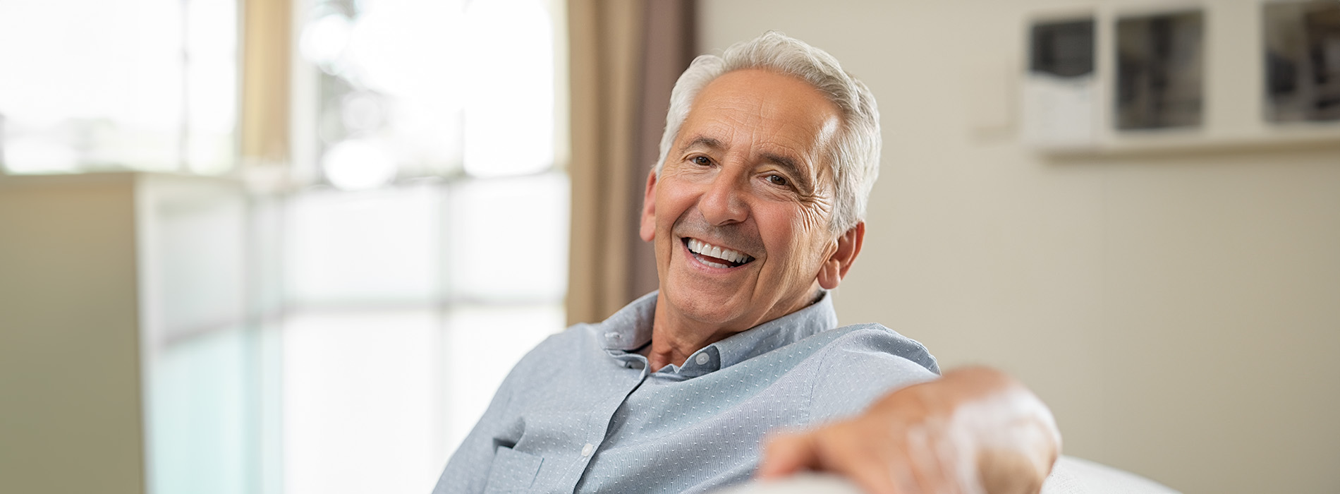 The image shows a man sitting in a room, smiling at the camera.