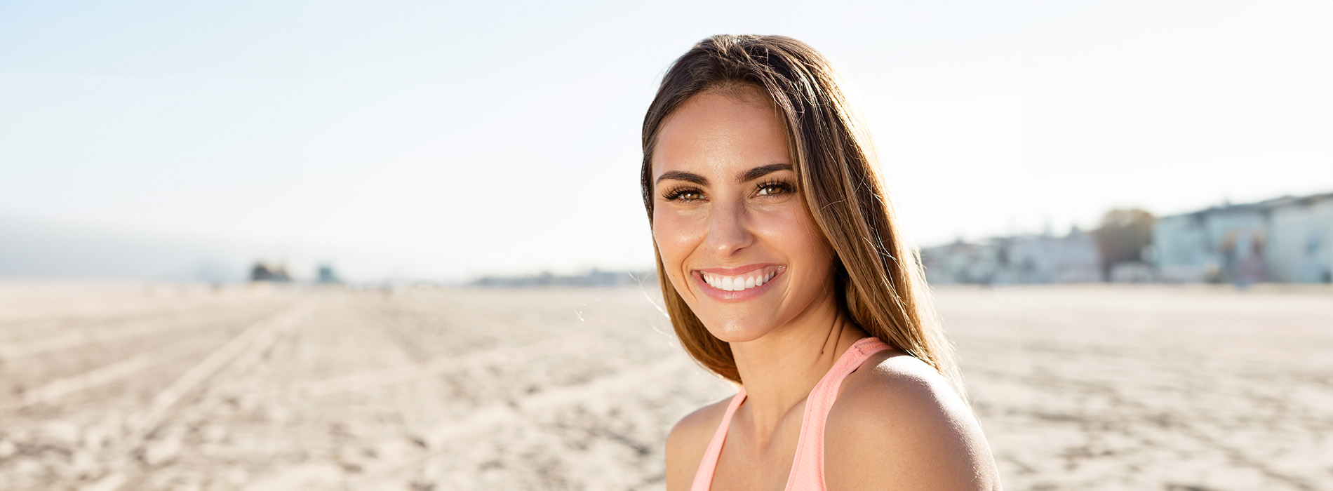 The image is a photograph of a woman standing on a beach with sand underfoot, wearing athletic attire and smiling towards the camera.