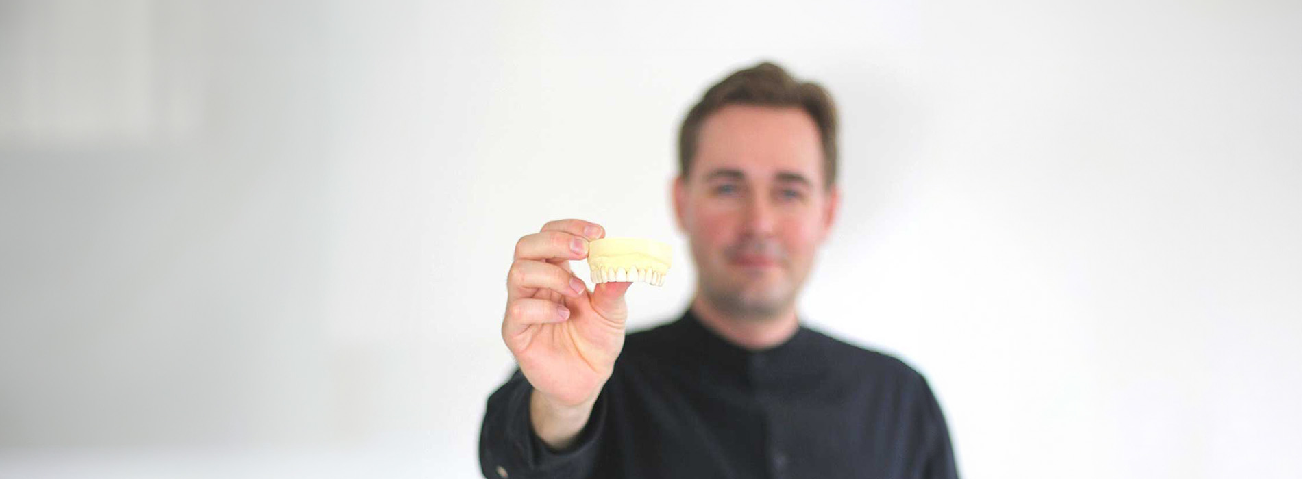 The image shows a man holding a small object, which appears to be a piece of paper or fabric, with his right hand extended towards the camera. He is standing against a plain background, and there is a visible reflection on the surface he is standing on.