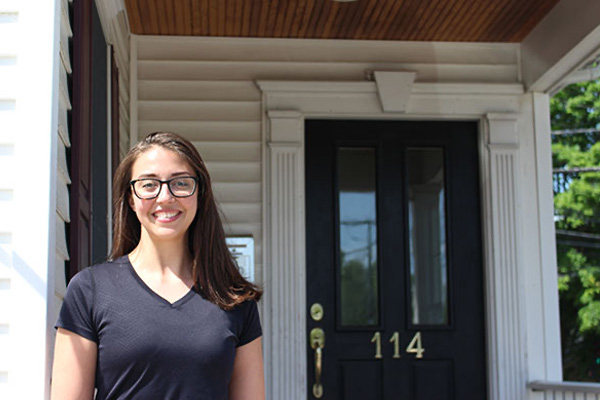 A woman in a black shirt stands in front of a white house with a black door, smiling at the camera.