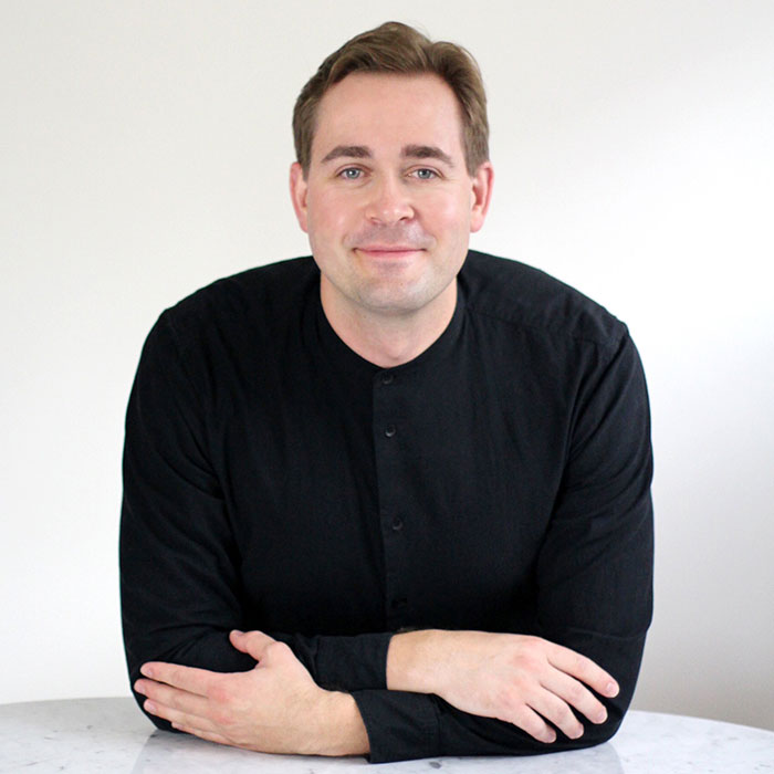 The image shows a man with short hair, wearing a dark shirt and standing in front of a white wall. He is leaning slightly to one side, resting his left hand on the table while looking directly at the camera with a neutral expression.