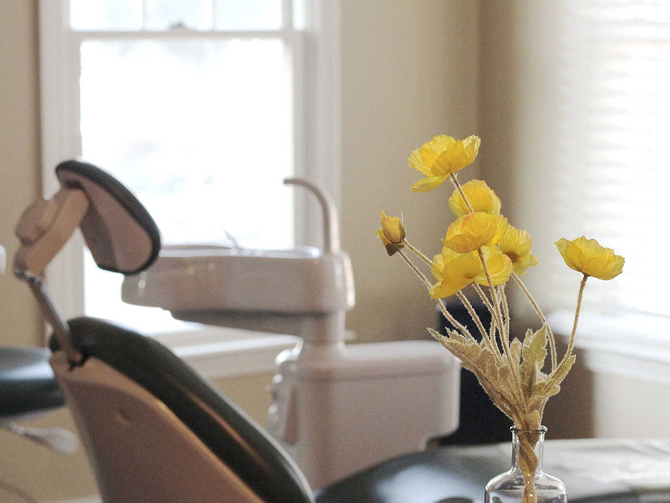 The image shows a dental office interior with a chair, desk, and various equipment, featuring a vase with yellow flowers on the right.