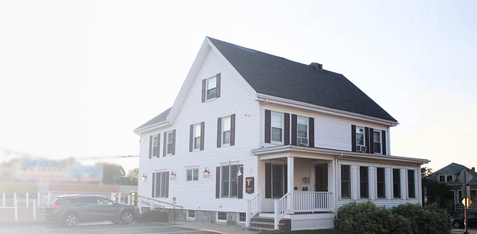 The image shows a two-story house with a black roof, white siding, and multiple windows. A small porch is visible at the front of the house, which appears to be a residential property during daylight.