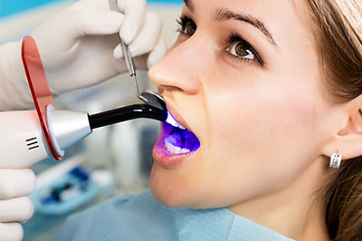 The image shows a woman sitting in a dental chair with a purple mouthpiece inserted, receiving dental treatment, while a dentist in a white coat and surgical mask is using a device to clean or examine her teeth.