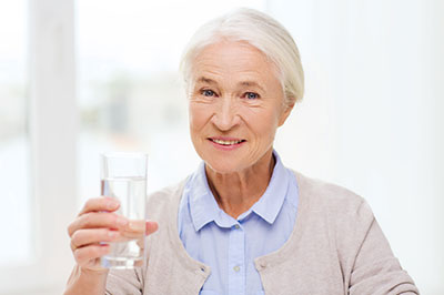 An elderly woman holding a glass of water, smiling and appearing to be in good health.