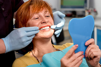 The image shows a woman seated in an orthodontist s chair, receiving dental care. She is holding up a blue model of her teeth and smiling at the camera. A dental professional is adjusting her braces with a pair of tweezers while wearing gloves and a face mask.