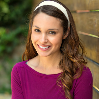 A woman with long brown hair wearing a purple top and a headband, smiling at the camera.