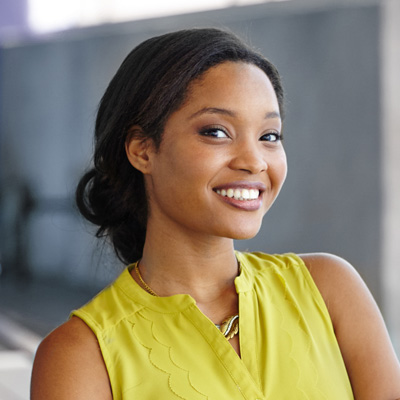 A smiling woman with dark hair, wearing a yellow top and standing against a concrete wall.