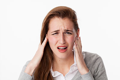 The image shows a woman with her hand on her head, appearing to be in pain or discomfort. She is wearing a grey top and has blonde hair.