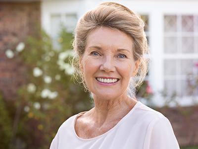 The image is of an older woman with blonde hair, wearing a white top and standing in front of a house.