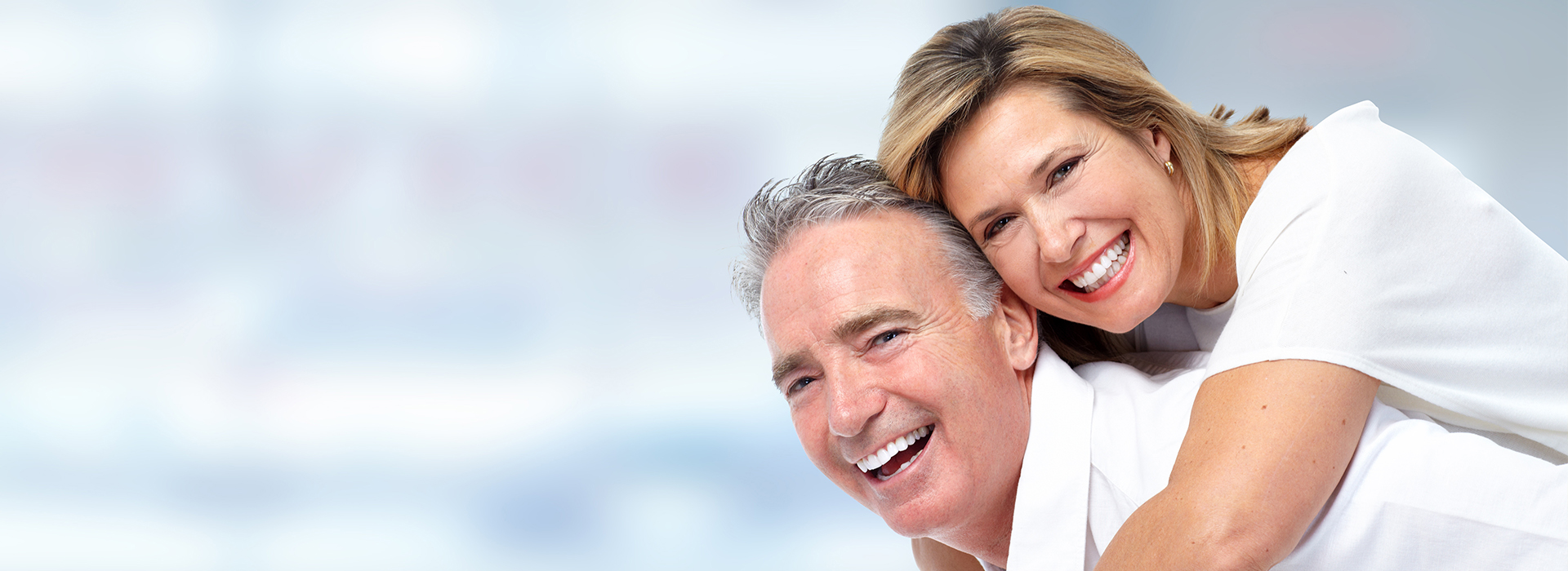 The image shows an older couple, a man and a woman, with the woman on the left smiling at the camera and the man on the right looking downward. They are both wearing white tops and appear to be embracing each other.