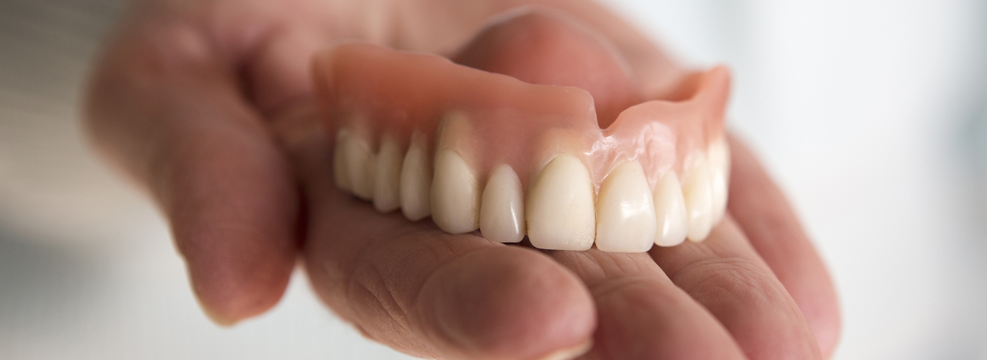 The image displays a person s hand holding a set of dentures against a blurred background.