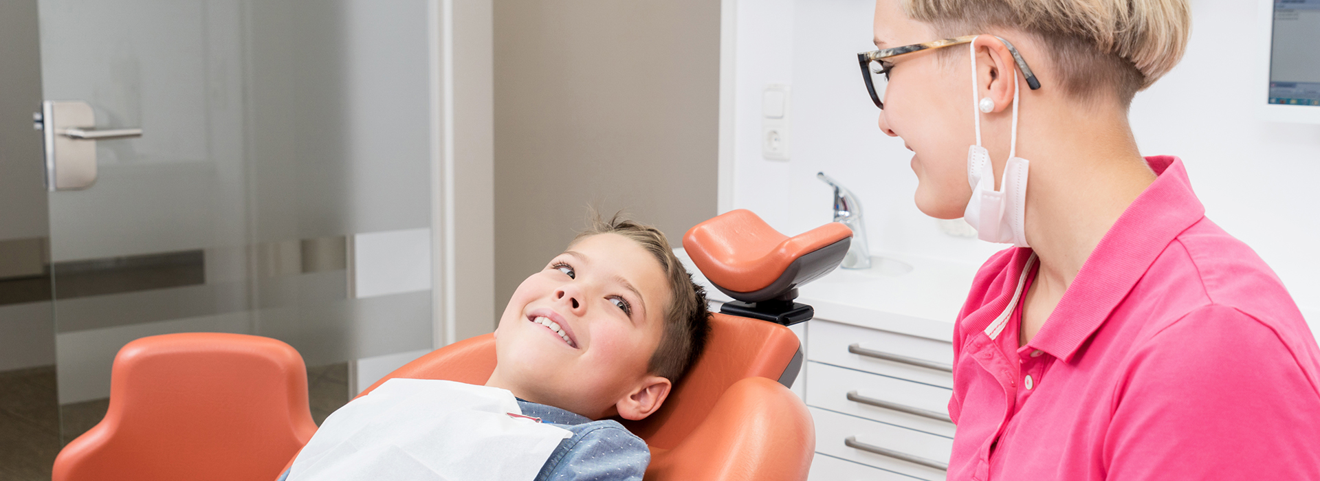 A young child sitting in a dental chair, smiling and looking at the camera, with a woman standing beside him.