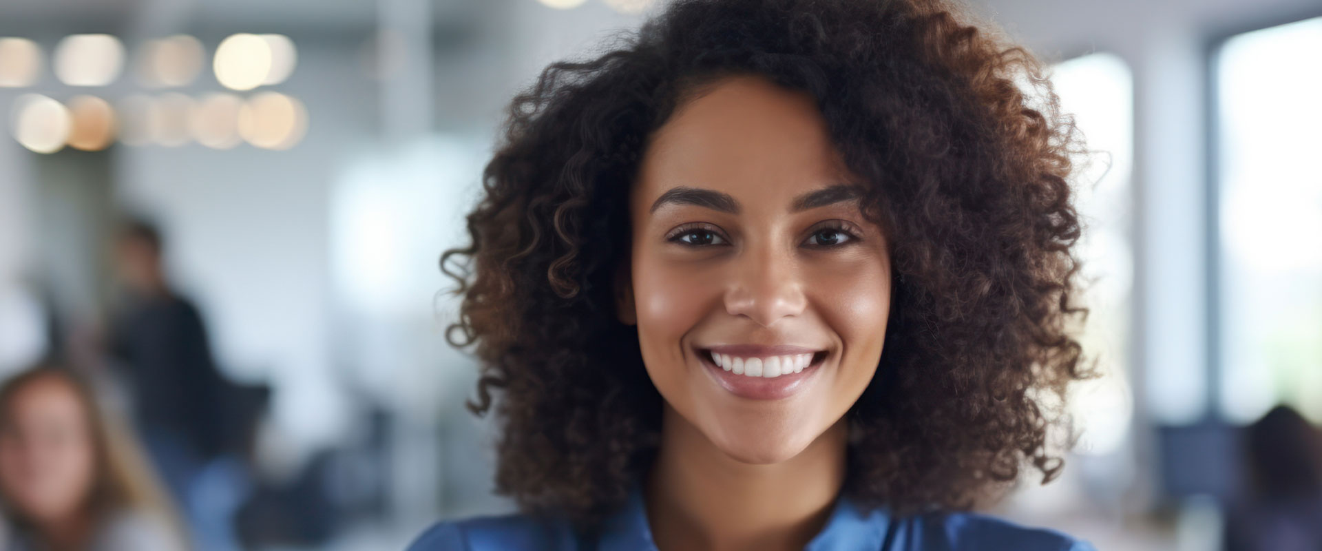 The image shows a person with curly hair smiling at the camera, wearing a blue top and standing in an indoor setting with a blurred background of other people.