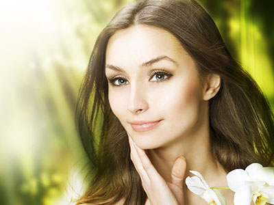 A woman with fair skin, light green eyes, and long hair is captured in a close-up portrait against a blurred background of greenery.