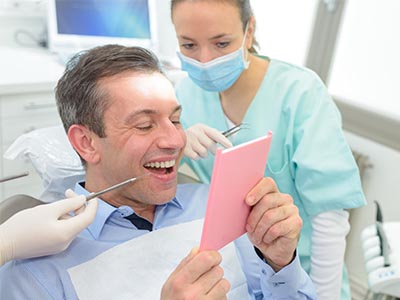 The image depicts a man sitting in a dental chair, holding a pink card or paper, with a smiling expression. Behind him, there s a dental professional wearing a surgical mask and holding a small tool, seemingly engaged in a conversation or examination with the seated man.