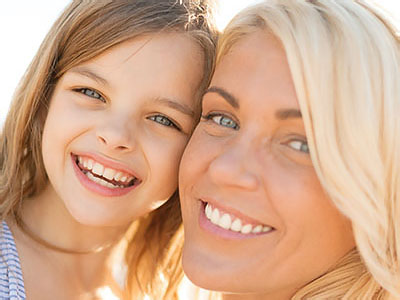 A smiling woman with blonde hair and a child in front of her, both outdoors on what appears to be a sunny day.