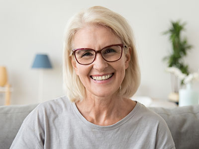 The image features a woman with blonde hair and glasses, smiling at the camera. She is wearing a light-colored top and has short hair. Behind her, there s a blurred background that suggests an indoor setting with home furnishings.