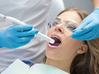 The image shows a woman in a dental chair receiving oral care, with a dental professional using an electric toothbrush on her teeth while she wears protective eyewear.