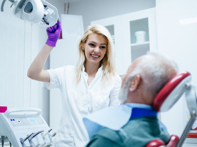 A dental professional in a white coat and gloves, smiling at the camera while standing next to an older patient with grey hair who is sitting in a dentist s chair with their mouth open.