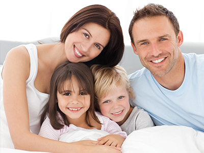 A family of four, including an adult couple and their two children, posing for a photo in a bedroom with a white bedspread.