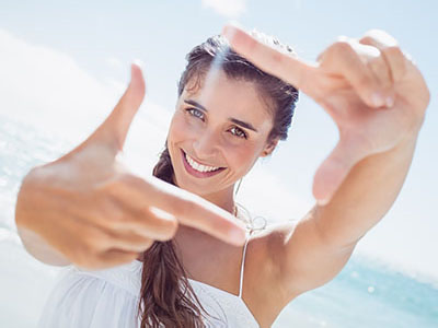 A woman with a radiant smile is taking a selfie, capturing her joyful moment on a sunny beach.